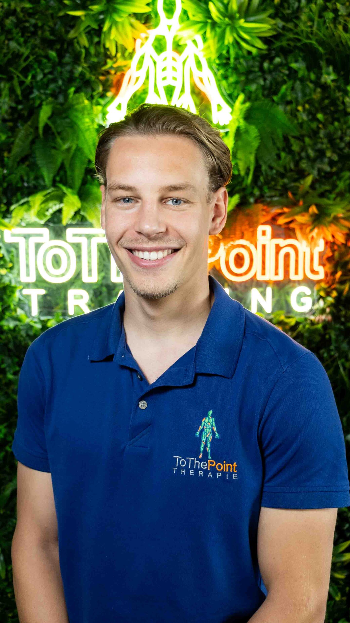 Fysiotherapeut Maxim bij ToThePoint Therapie poseert voor de groene wand met neonlogo.