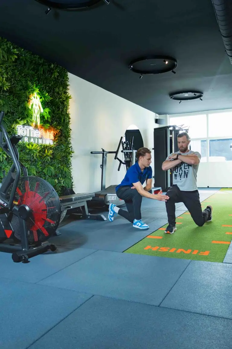 Personal trainer begeleidt een klant tijdens een oefening in een moderne gym, met een groene wand en fitnessapparatuur op de achtergrond.